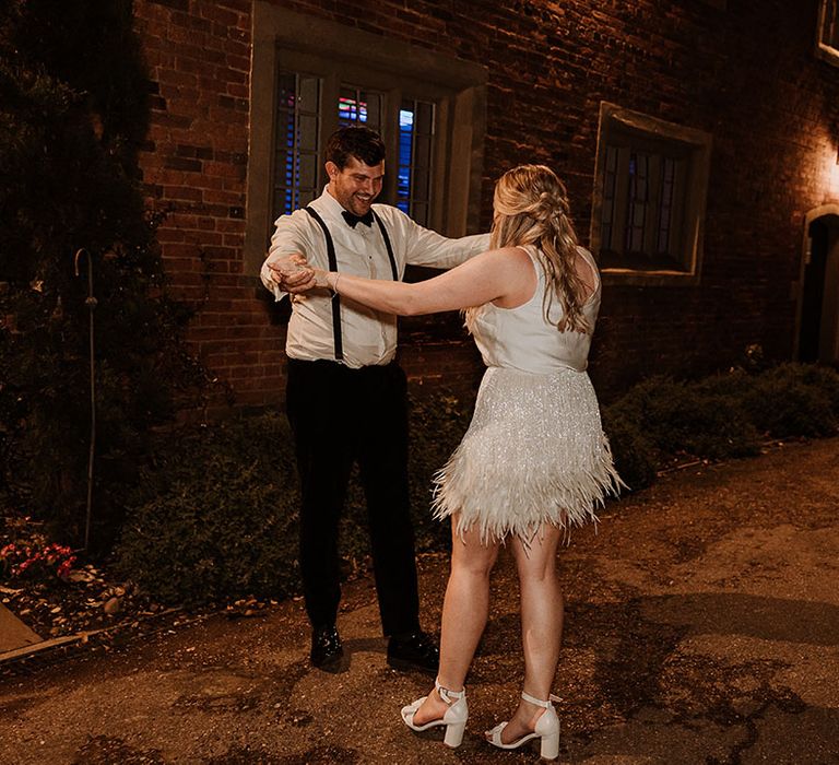 Bride in block heel white wedding shoes in sparkly tassel skirt and sleeveless white top with groom in white shirt and black trousers 