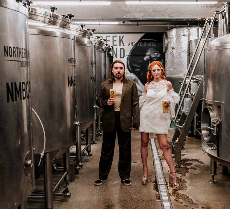 Groom in brown blazer, white shirt, grey suit trousers and vans shoes standing with bride in 70s-inspired boatneck short lace wedding dress with detachable tulle puff sleeves and platform gold wedding heels holding pints of beer by beer fermenters