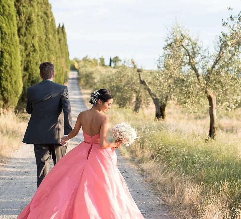 Bride in strapless pink wedding dress with white floral bouquet walking hand in hand with the groom 