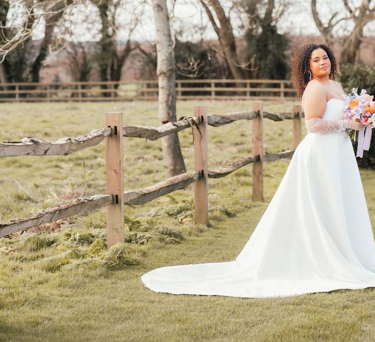 Bride in strapless wedding dress with front slit, puddle train and mesh pearl gloves holding orange, lilac and peach mixed flower bridal bouquet 