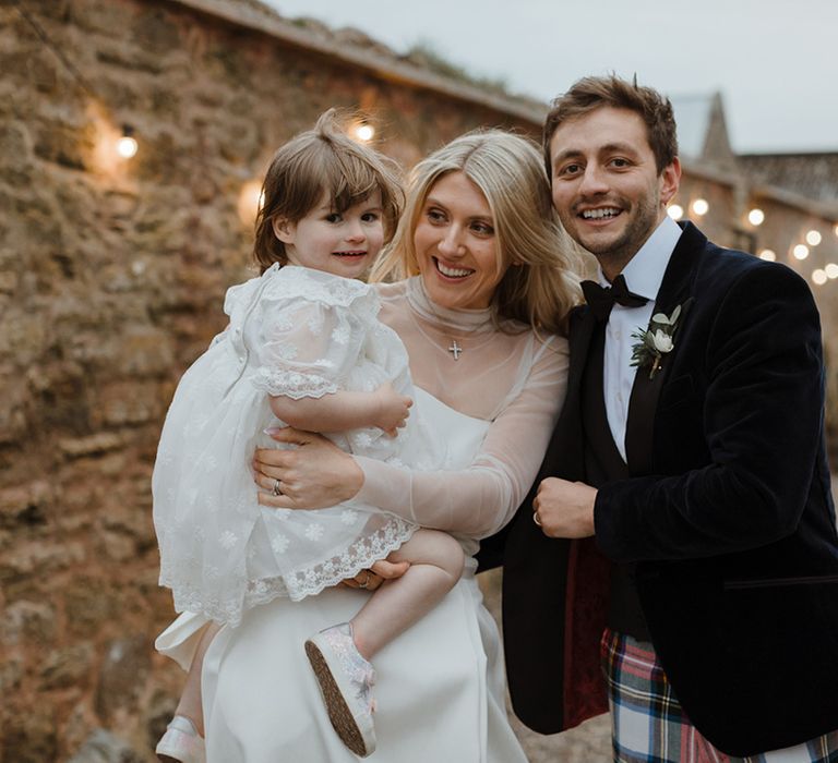 The bride and groom carry a flower girl in a white dress with white faux fur cardigan 