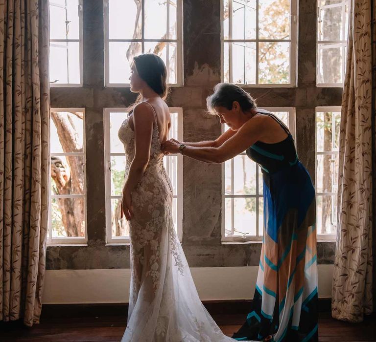 Mother of the bride in tropical multicoloured summer dress helping bride into her lace sleeveless wedding dress with puddle train