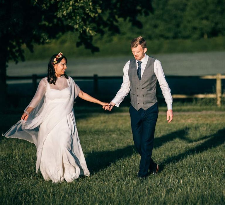 Groom in grey waistcoat walking hand in hand with bride in long sleeve mesh overlay wedding dress and colourful bridal flower crown on the grounds of Templars Barn