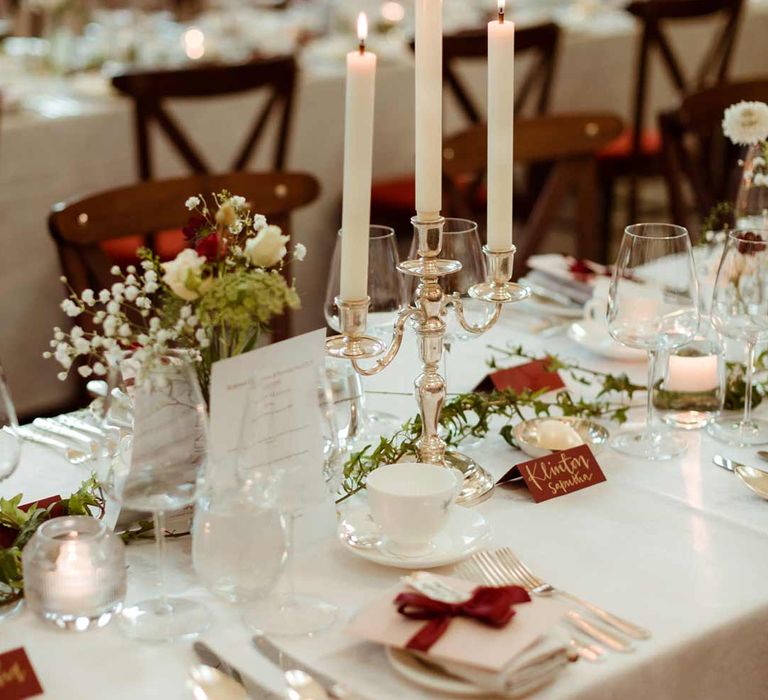 Classic wedding tablescape with white tapered candles in gold candelabras, red wedding table names, white rose, baby's-breath and foliage wedding centrepieces and foliage table runners