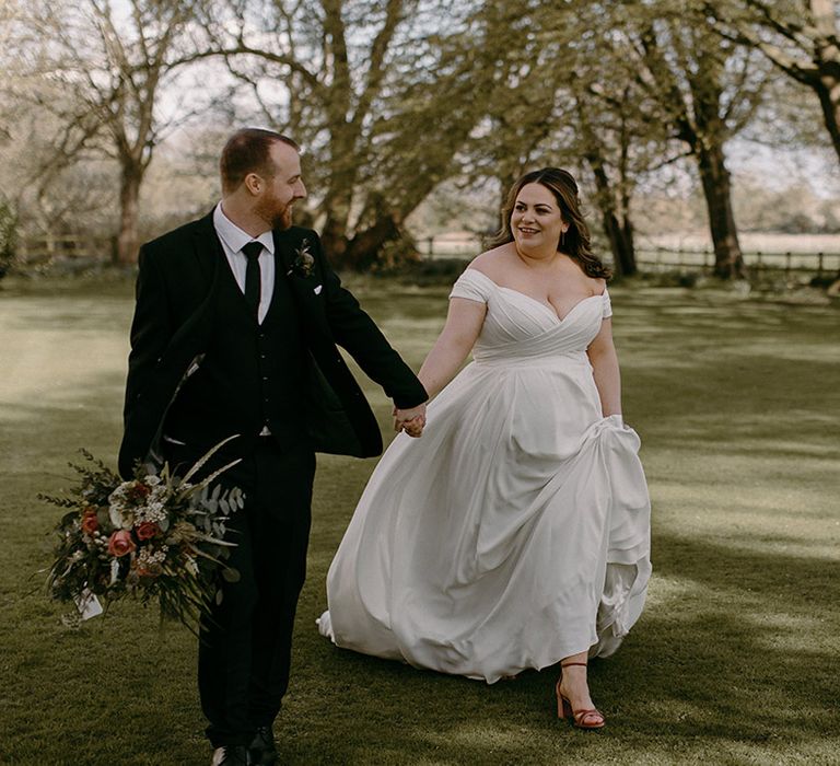 Groom in black and white traditional wedding walking hand in hand with the bride in off the shoulder Madi Lane wedding dress 