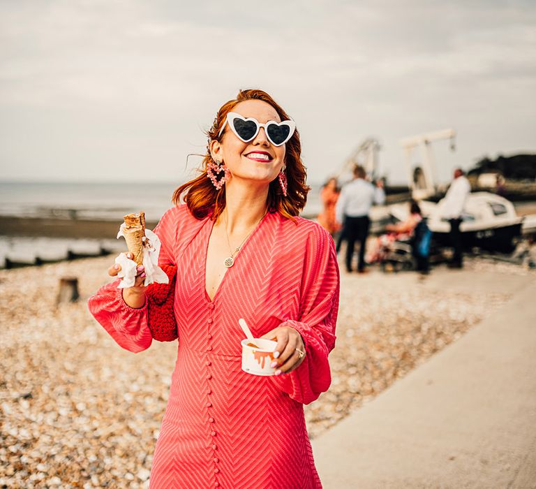 guest in a red midi wedding guest dress earring white heart sunglasses and eating ice cream at coastal wedding 