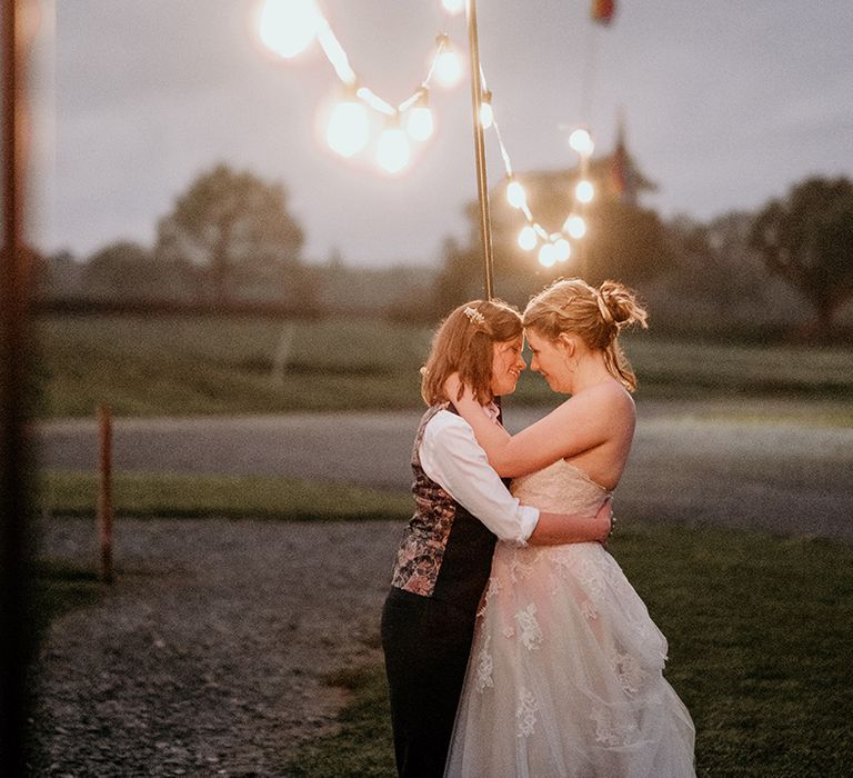 two brides in a waistcoat and lace wedding dress embracing under a canopy of festoon lights 