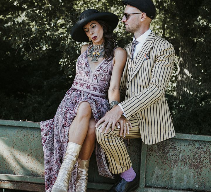 Bride wearing a pink boho wedding dress with silver wedding boots, aztec wedding jewellery and a boho bridal hat sits next to groom wearing a mustard pinstripe suit, lilac socks, black sunglasses and a black hat