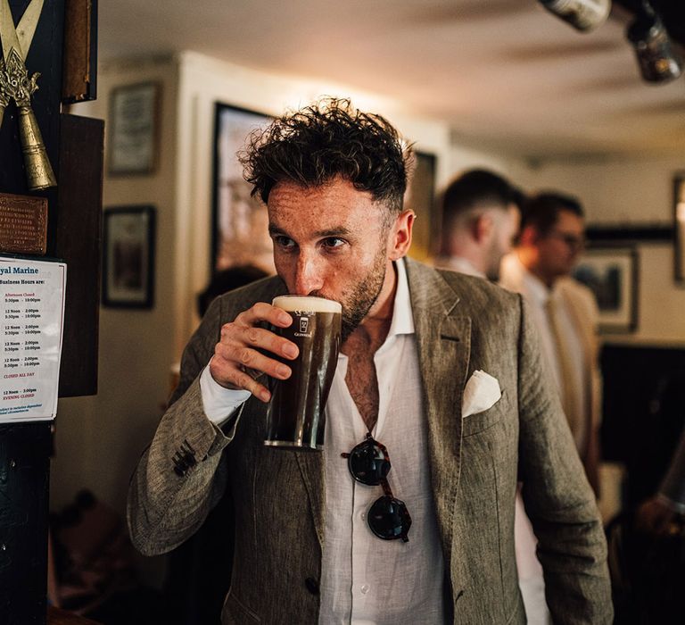 Groom wearing white linen shirt, grey suit jacket drinking pint go Guinness in a local pub before the wedding 