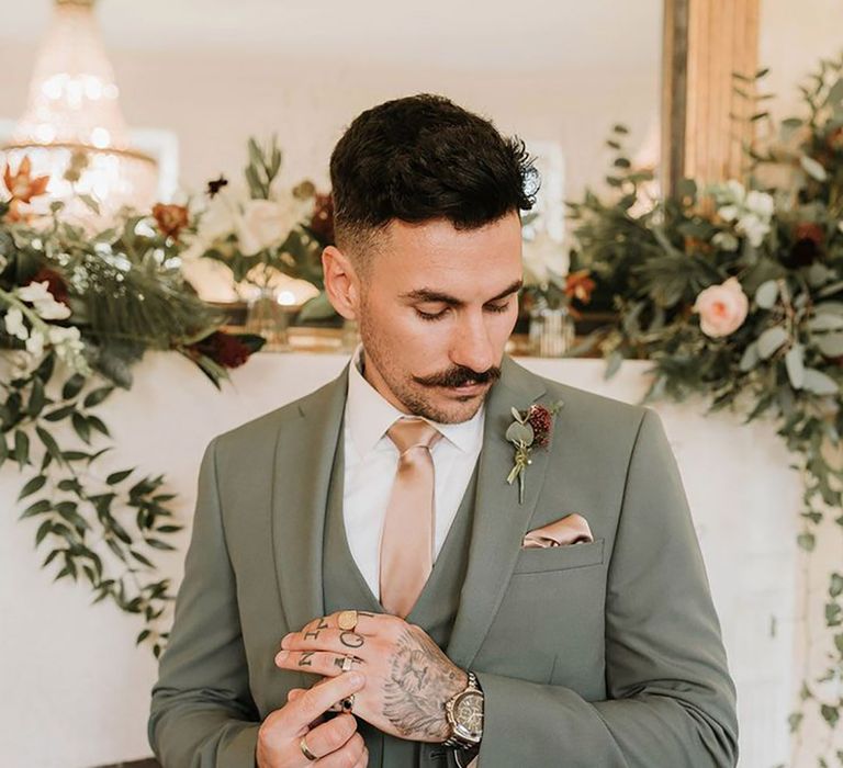 Groom at Christmas wedding wearing a sage green wedding suit with satin tie and pocket square with flower buttonhole 