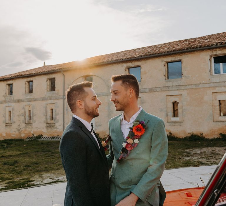 Two grooms at their same sex wedding next to a red vintage wedding car 