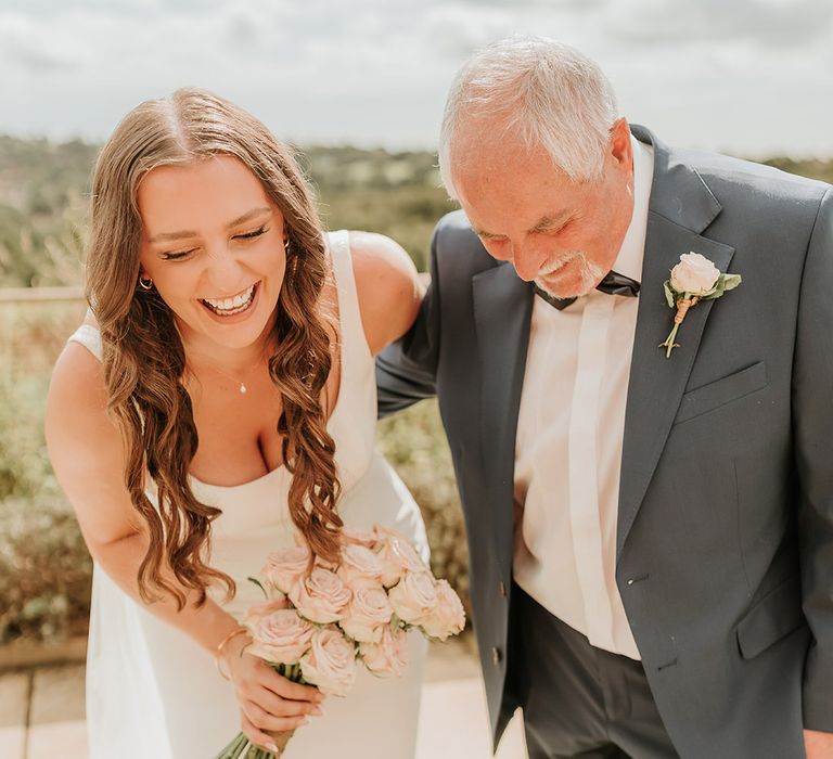 Father of the bride in blue wedding suit laughs with the bride in a satin slip wedding dress for their first look moment