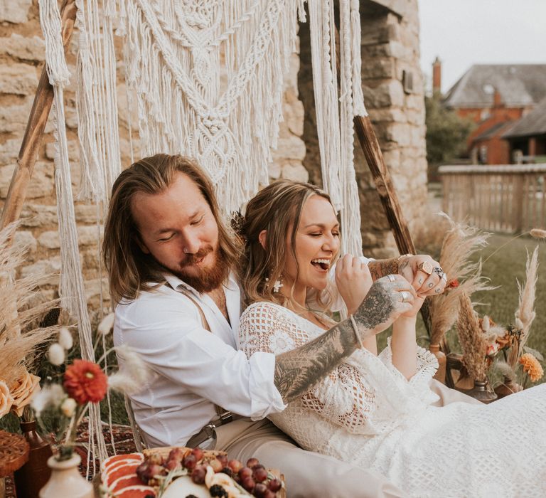 Bride & groom sit in front of Macrame backdrop for outdoor picnic