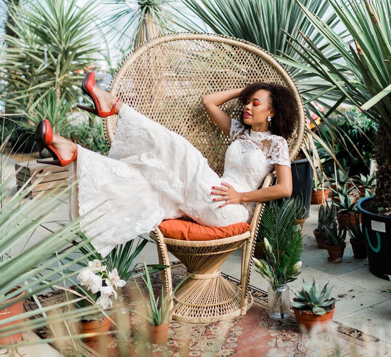 Bride in white Lillian West bridal jumpsuit and red Next heels sits on rattan throne