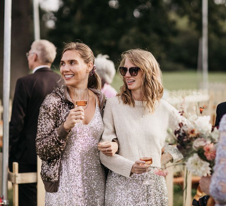 Bridesmaids in silver sequin dresses, bomber jacket and wool jumper drinking in a field 