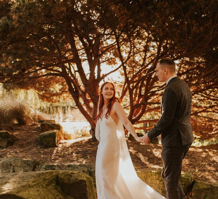 Bride and groom photography surrounded by trees and autumnal colours. Bride has red hair and simple silk wedding dress. Groom wears tailored check suit 