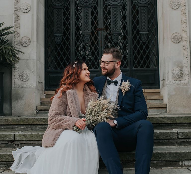 Bride in fur coat holding dried flower bouquet sits on steps with groom in navy suit and tartan waistcoat