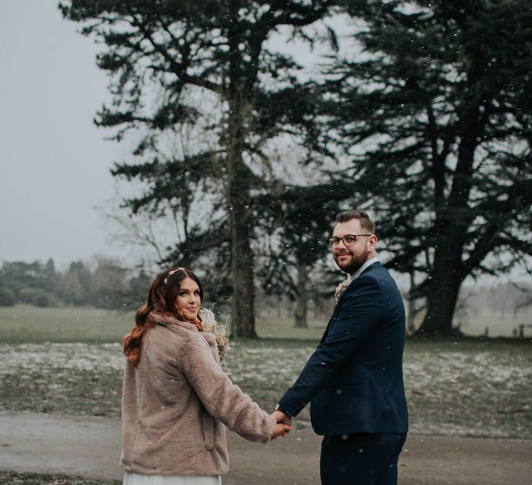 Bride in fur coat stands holding hands with groom in navy suit in the snow at Cannon Hall 