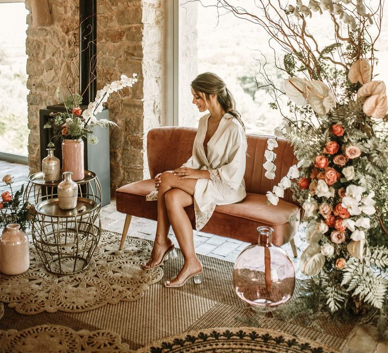 Bride on the wedding morning in a sating getting ready robe sitting on a pink velvet love seat surrounded by boho wedding flowers