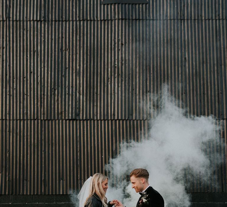 Bride in personalised leather bridal jacket and white Charlie Brear wedding dress holding hands with groom in black Hugo Boss suit with white smoke bomb in background