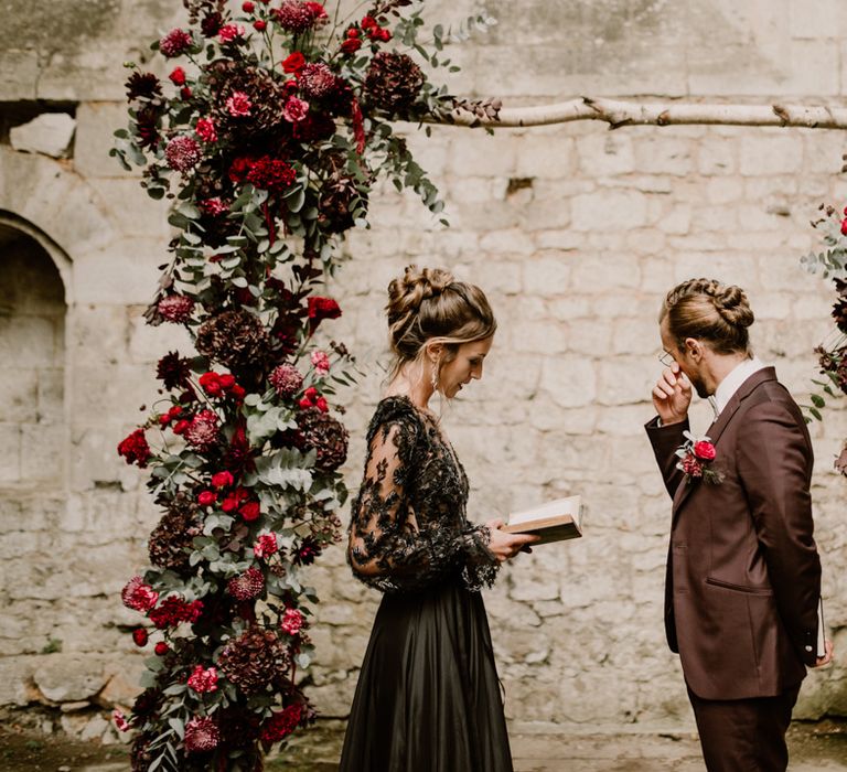 Emotional groom in a burgundy suit standing at the wooden altar decorated with red flowers with his bride in a black wedding dress reading her vows 