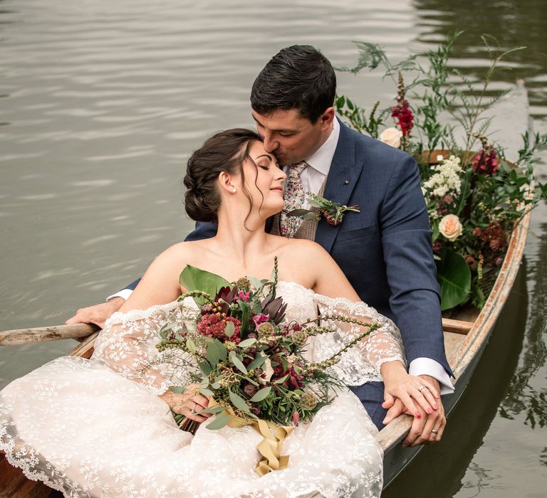 Bride & groom sit in boat as it floats along the river whilst bride holds bouquet