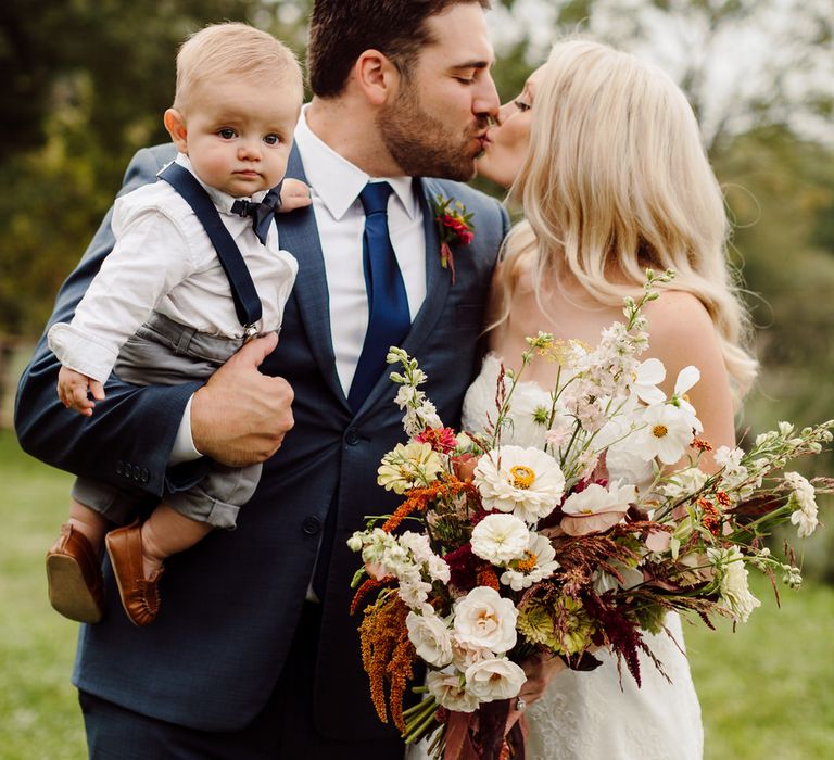 The bride and groom kiss while the groom holds their 7 month old son, who looks at the camera