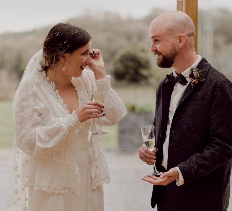 Bride in white blouse over Charlie Brear wedding dress wearing applique veil wipes tear from her eye whilst smiling at emotional groom in grey tweed suit