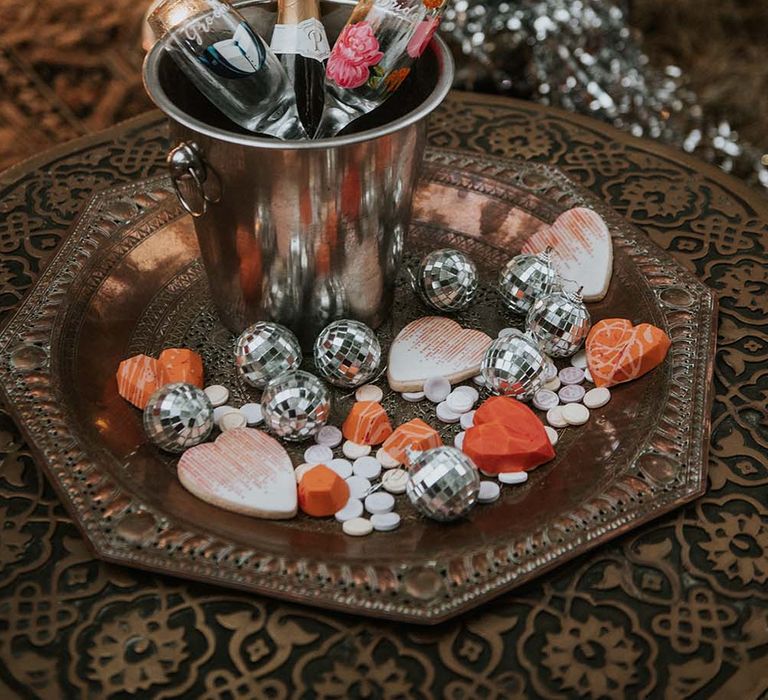 Prosecco bottle and glasses in ice bucket on bronze octagonal tray with mini disco ball and heart decor at festival themed wedding