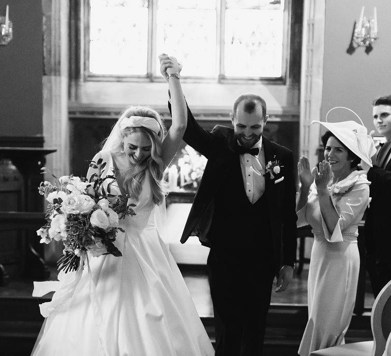 Black & white image of bride & groom celebrating after wedding ceremony as they lift their hands together in celebration and wedding party claps