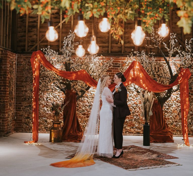 Two brides hugging in front of indoor blossom trees for pumpkin spice wedding theme