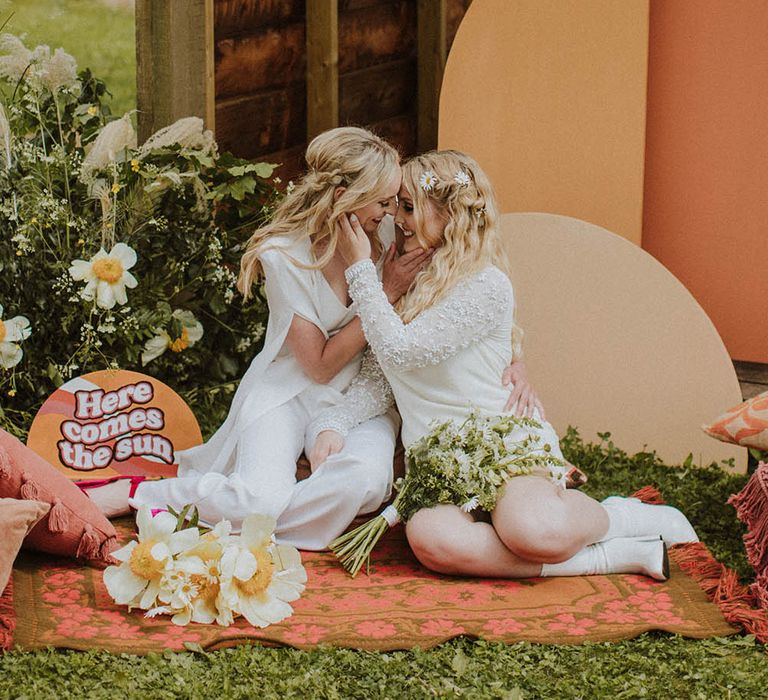 Two brides sitting on a wool rug with retro 'Here Comes The Sun' wedding sign and colour block altar decor 