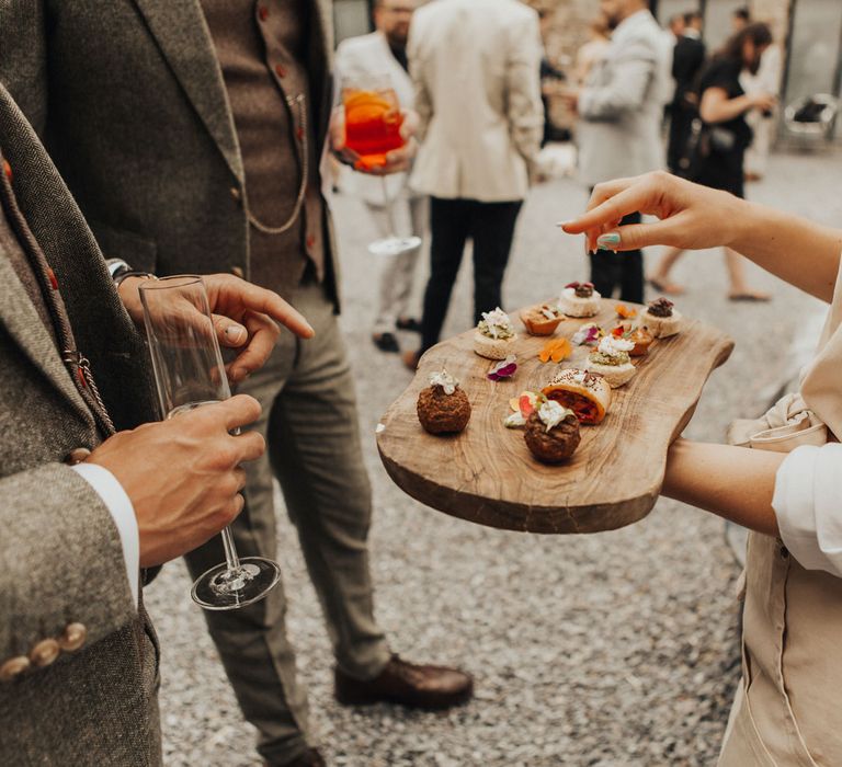 Waitress holds wooden board with assorted canapés on as guests in sage green suits holding drinks have a look in courtyard during wedding reception at Anran Devon