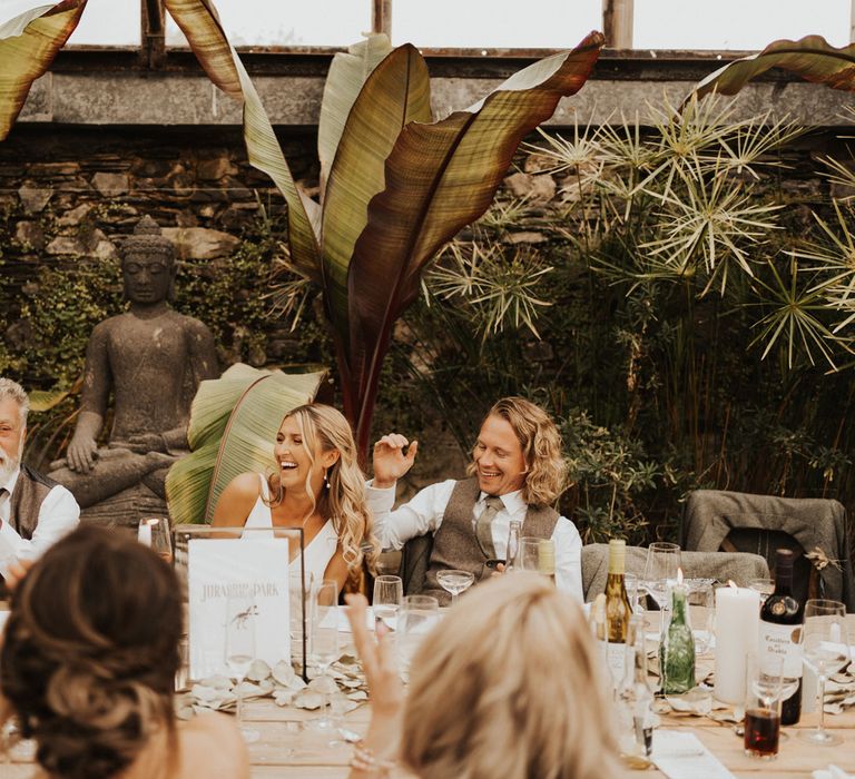 Bride in white Made With Love wedding dress laughs with groom in sage green waistcoat and tie and guests as they listen to speeches whilst sat at wooden table in the glasshouse at Anran Devon during wedding breakfast