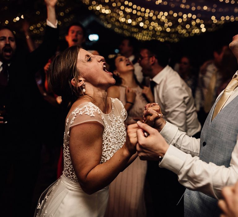 Bride in Rime Arodaky wedding dress holds hands with groom in blue waistcoat and gold tie under a canopy of fairy lights at evening wedding reception as they both sing