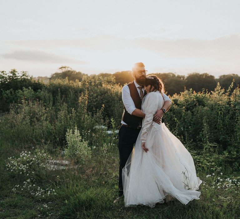 Bride & groom stand within field on their wedding day as the sun goes down 