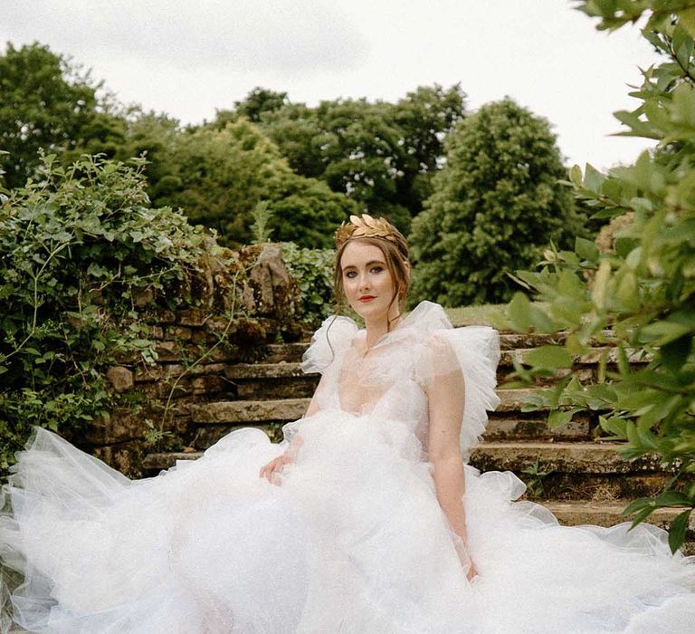 Beautiful bride in a tulle wedding dress with embellished wedding shoes wearing a gold crown sitting on the steps at Bourton Hall 