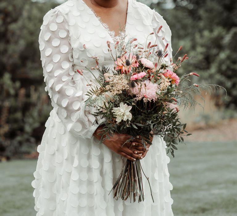 Bride in long mesh sleeve spotted Vagabond wedding dress with bridal hair bow holds pink, green and white mixed bridal bouquet at Wasing Park wedding