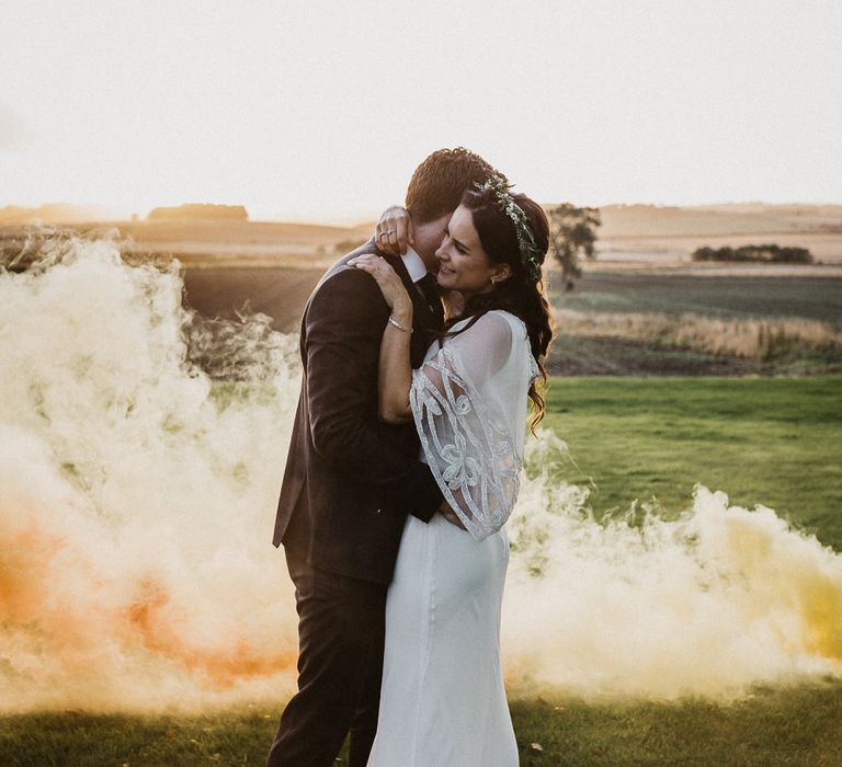 Bride in an ethereal wedding dress with lace sleeve design embracing her husband in a field with an orange smoke bomb blowing in the background 