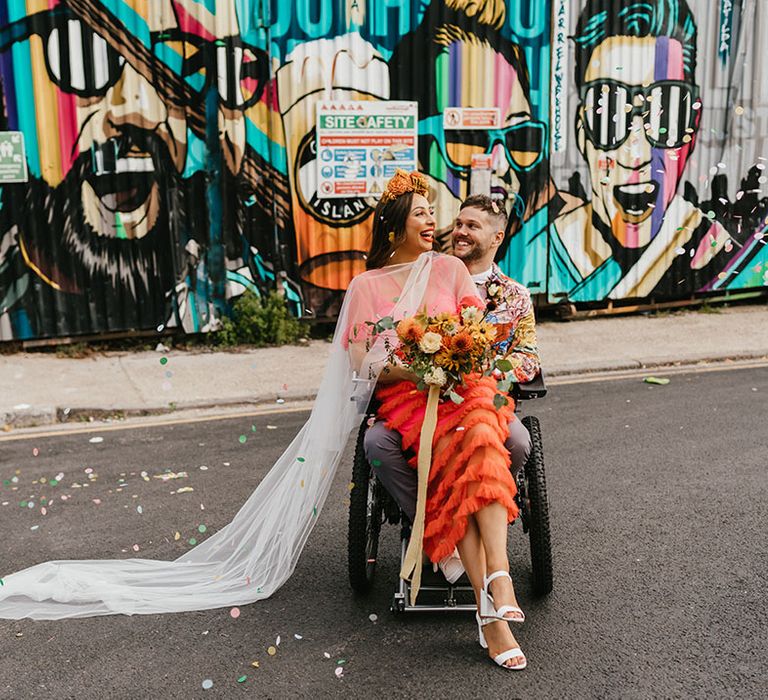 Bride in a coral wedding dress with embroidered veil sitting on her grooms lap in a wheelchair in Shoreditch 