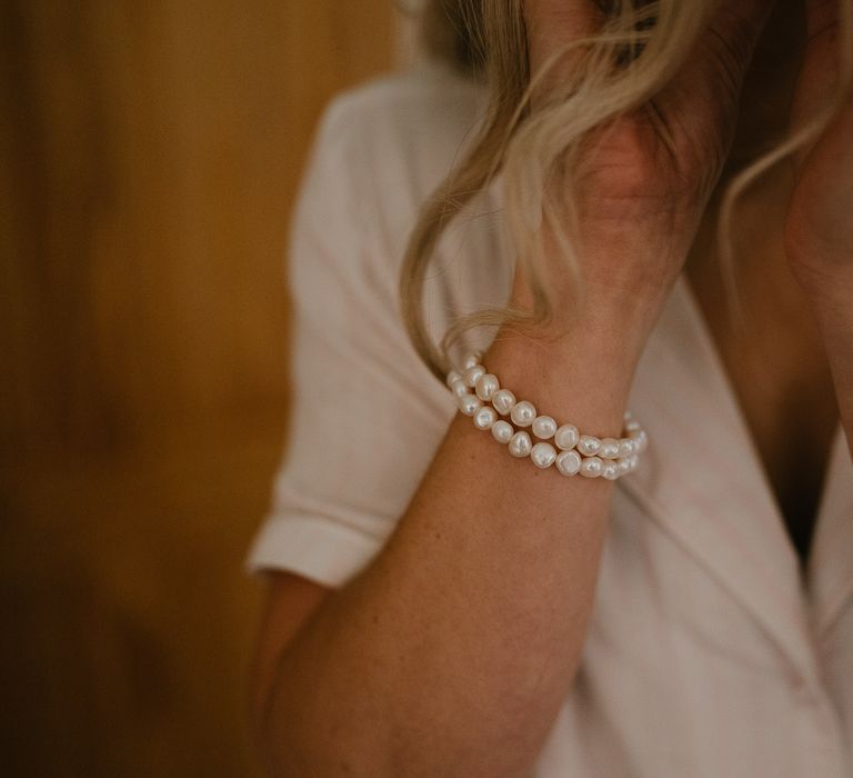 Bride with blonde curled wedding hair wearing two string pearl bracelet puts earrings in before late summer wedding in Norfolk