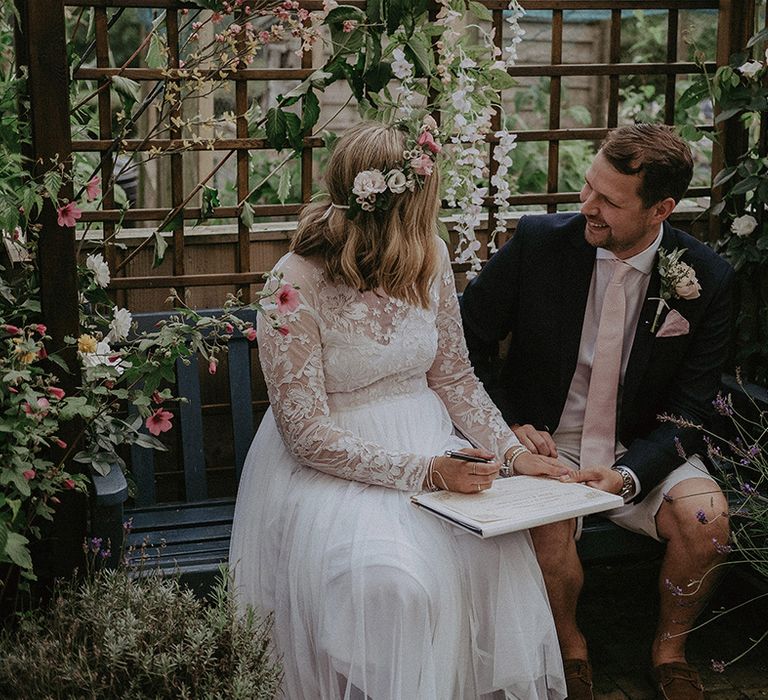 Bride in a floaty wedding dress with lace sleeves and flower crown sitting on a bench with her husband in beige shorts and a navy blazer at their back garden wedding 