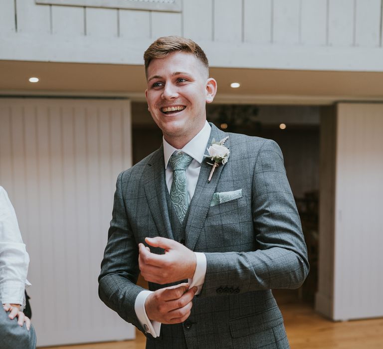 Groom in grey checked three piece suit with mint green paisley tie and pocket square and floral buttonhole adjusts cuffs before summer wedding at Primrose Hill Farm