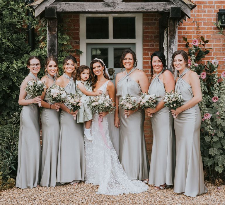 Bride in Berta wedding dress with applique flowers holding flower girl in sage green dress stands in front of farmhouse with bridesmaids insane green halter neck bridesmaid dresses all holding white, blush pink and green wedding bouquets for summer wedding at Primrose Hill Farm