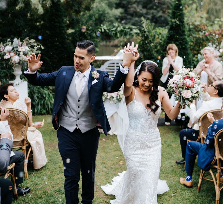 South Asian Bride in embellished detailed long fitted dress & South asian Groom in simple navy suit with a grey waistcoat happy as they pass their guests in simple yet stunning outdoor ceremony 