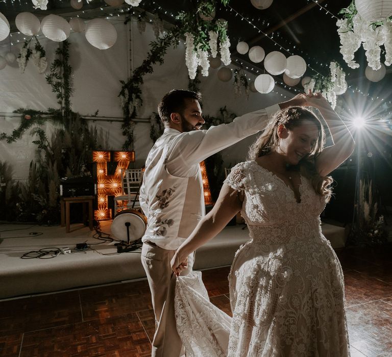 Bride in lace homemade wedding dress with train is twirled around dance floor by groom in beige waistcoat and suit trousers inside wedding reception marquee with garlands, paper lanterns and fairy lights at garden wedding in Bedfordshire