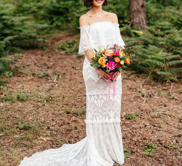 Bride in off the shoulder Grace Loves Lace wedding dress and pink and orange flower crown stands in woodland holding colourful bridal bouquet after woodland wedding ceremony at The Bridal Barn