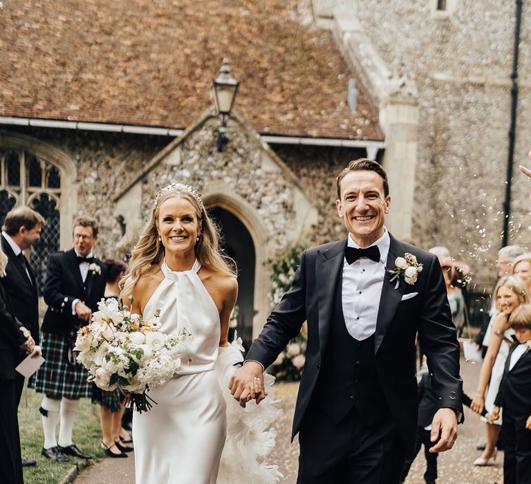 Bride in satin halterneck Halfpenny London wedding dress holding white and green wedding bouquet holds hands with groom in black tie as they walk through churchyard whilst guests throw confetti