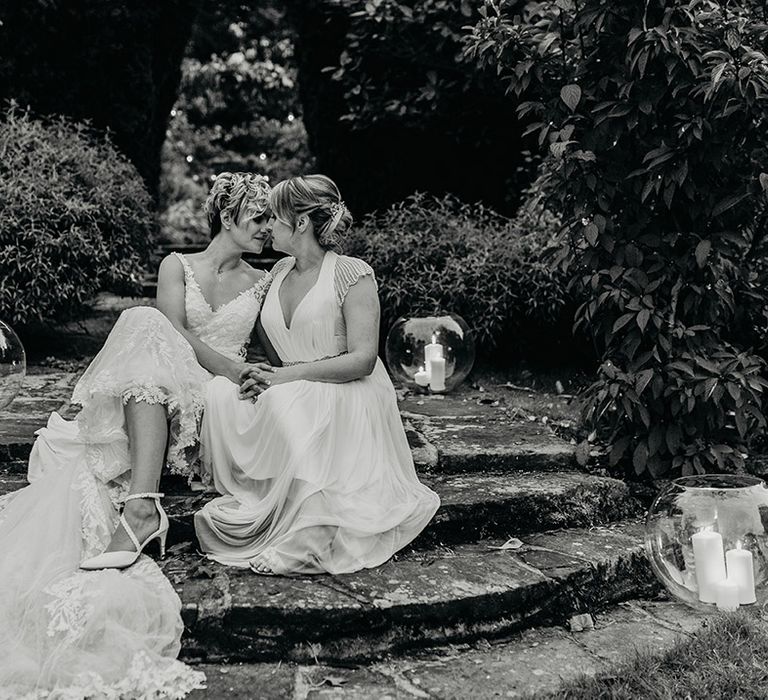 Black & white image of brides sitting on stairs outdoors surrounded by candles