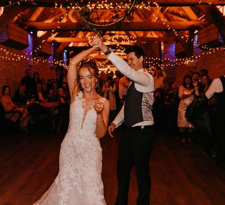 Groom in white shirt and dark waistcoat twirls bride in lace wedding dress around on the dance floor during rustic barn wedding reception lit by fairy lights